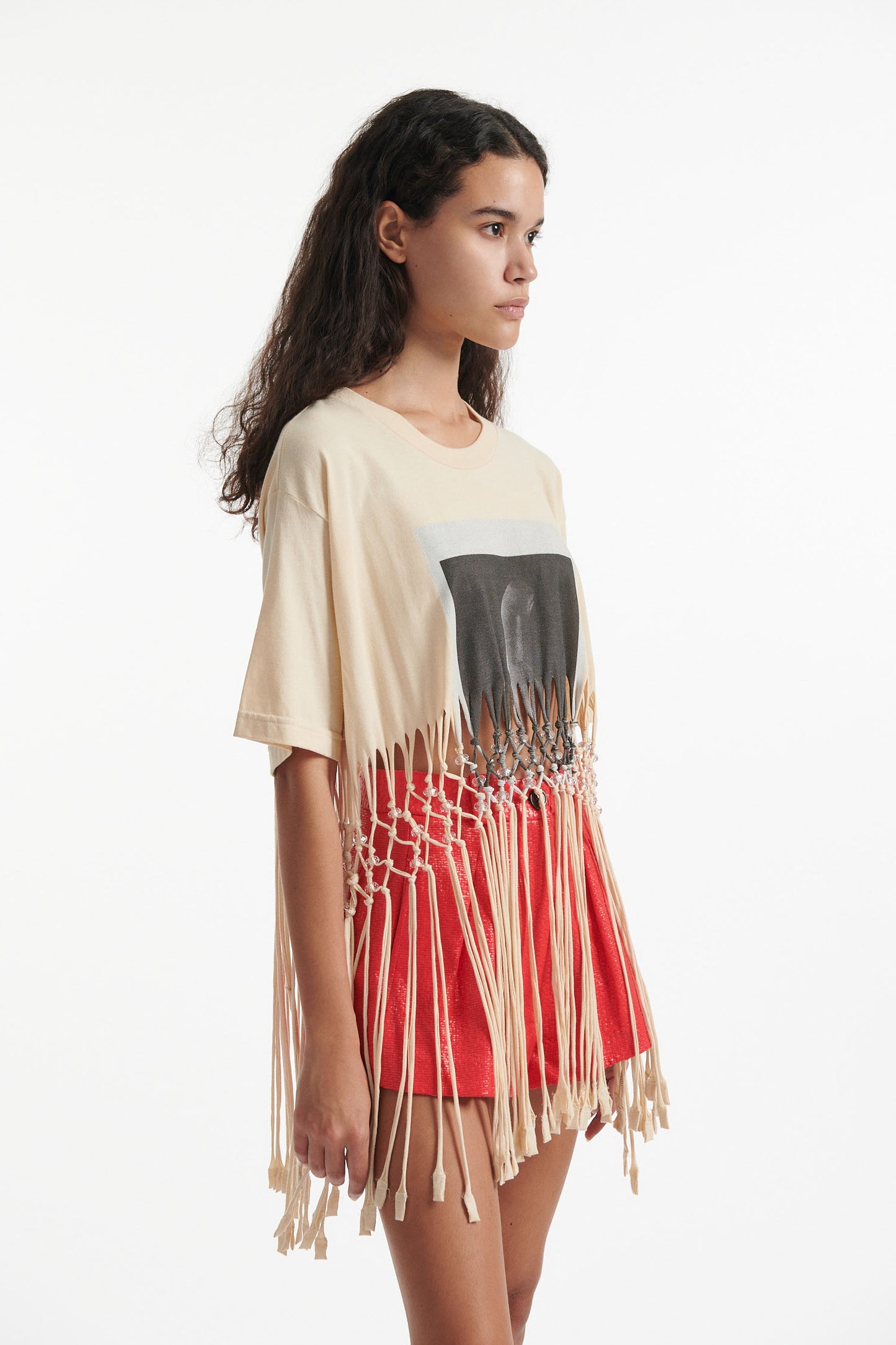 Female model wears deconstructed beige graphic tee with crystal embellishments and red shorts and black high heels on white background. 