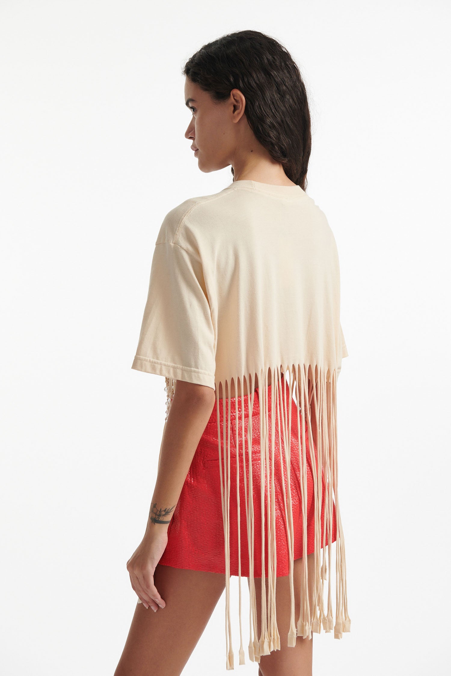 Female model wears deconstructed beige graphic tee with crystal embellishments and red shorts and black high heels on white background. 