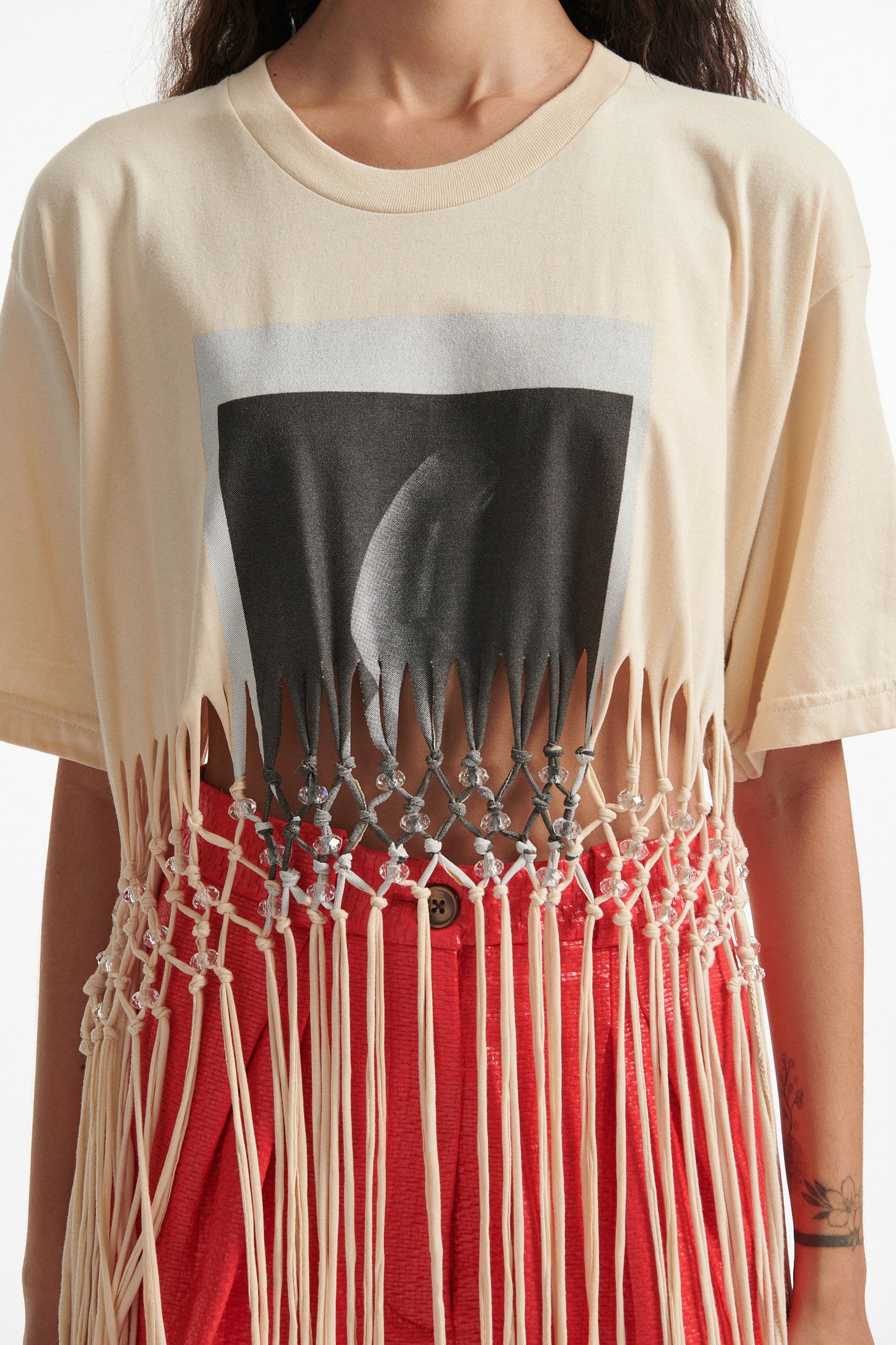 Female model wears deconstructed beige graphic tee with crystal embellishments and red shorts and black high heels on white background. 