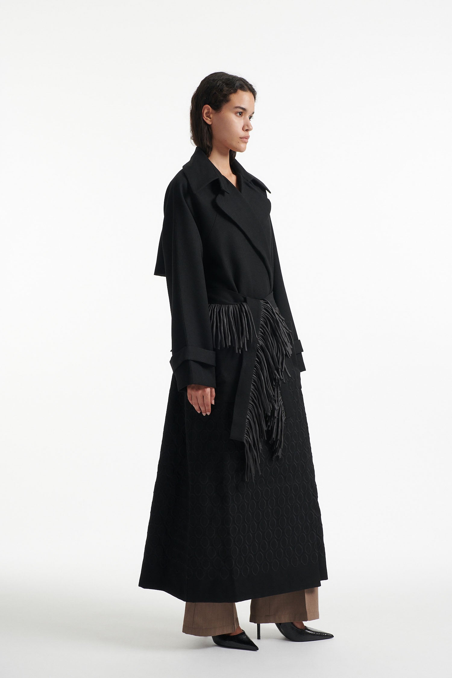 Female model wears black double breasted wool coat with fringe waist belt  and diamond embroidery with black high heel boots on white background. 