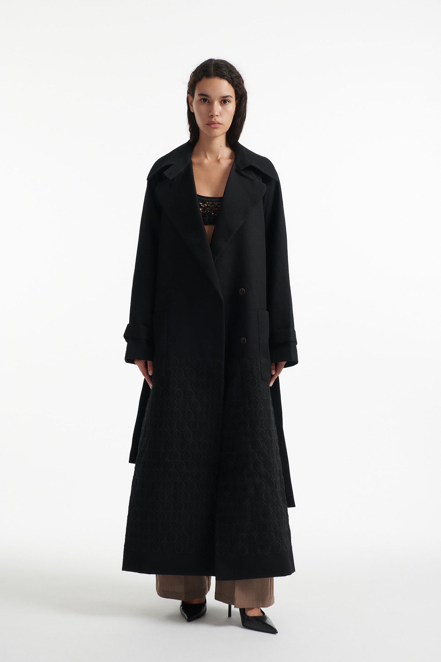 Female model wears black double breasted wool coat with fringe waist belt  and diamond embroidery with black high heel boots on white background. 