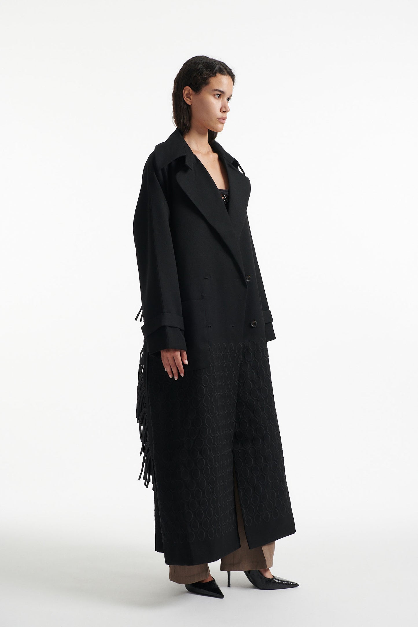 Female model wears black double breasted wool coat with fringe waist belt  and diamond embroidery with black high heel boots on white background. 