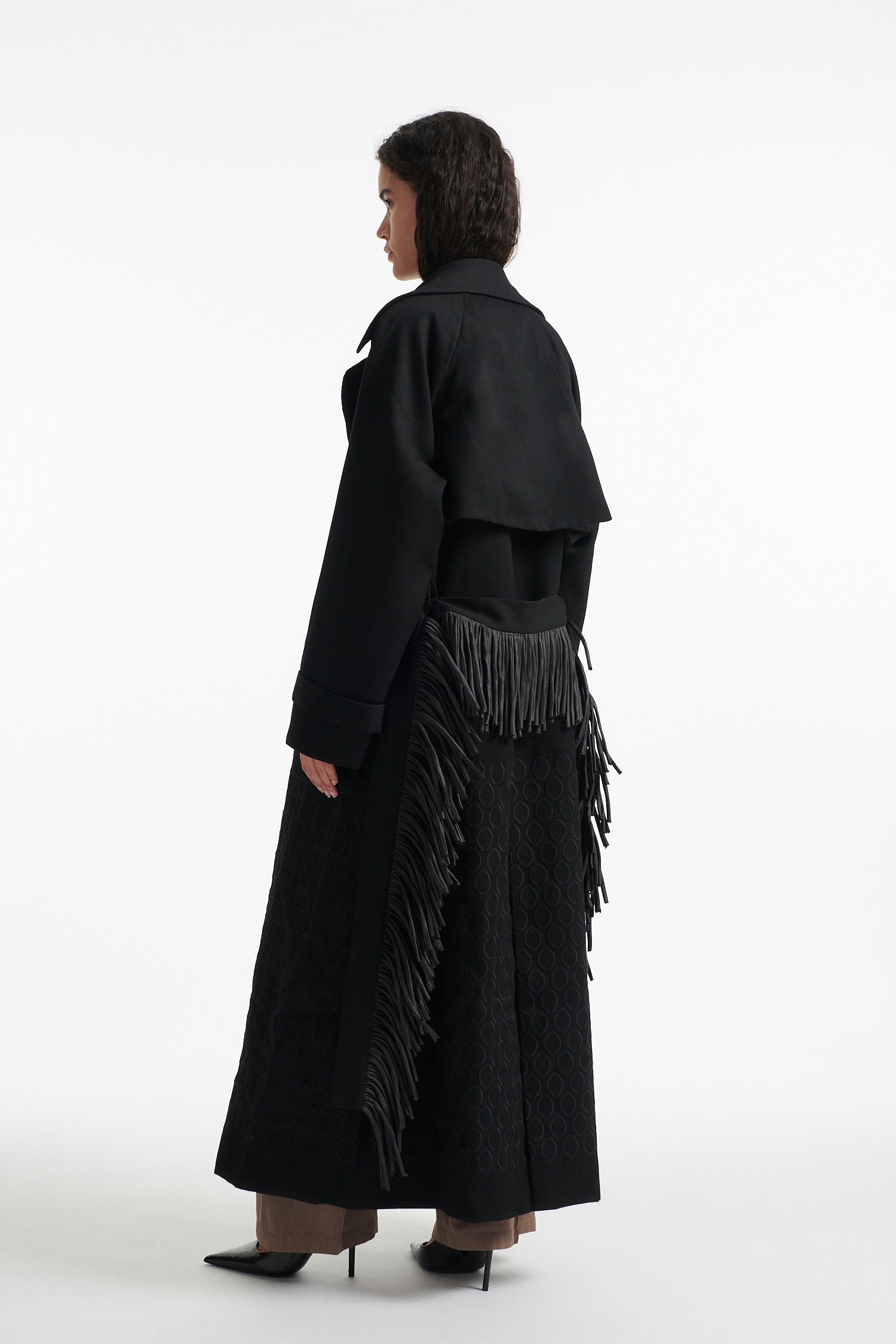 Female model wears black double breasted wool coat with fringe waist belt  and diamond embroidery with black high heel boots on white background. 