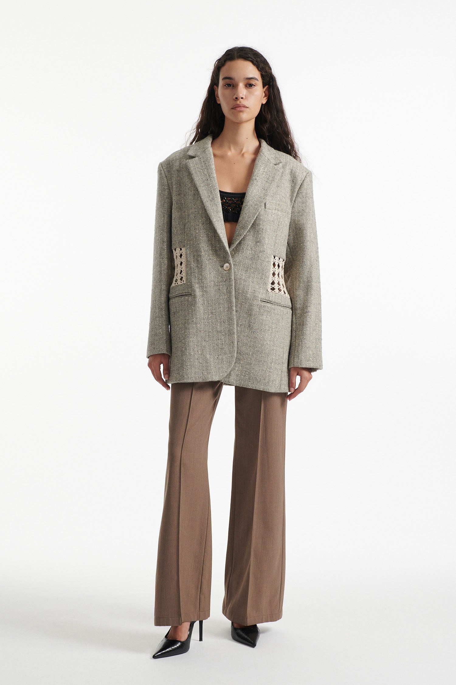 Female model wears light grey wool blazer with crochet paneling, brown crochet embellished trousers and black high heels on white background. 