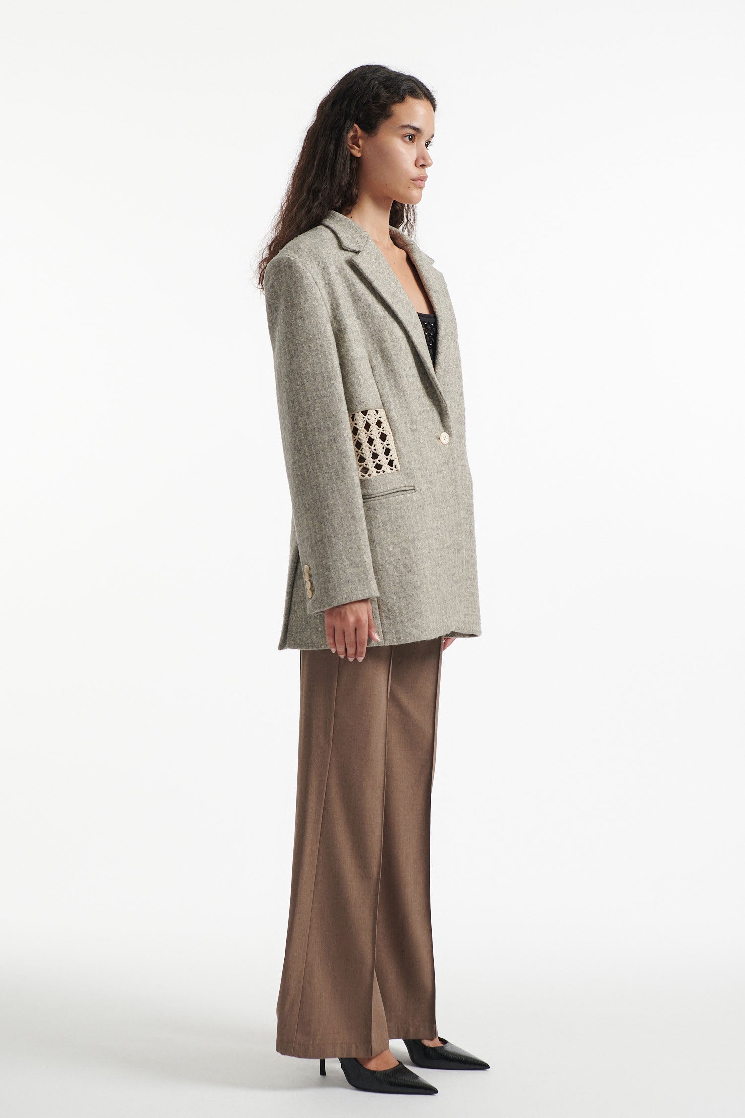 Female model wears light grey wool blazer with crochet paneling, brown crochet embellished trousers and black high heels on white background. 