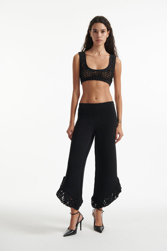 Female model wears short black knit trousers with fringe loops and black crystal embellished bralette with black high heels on white background.