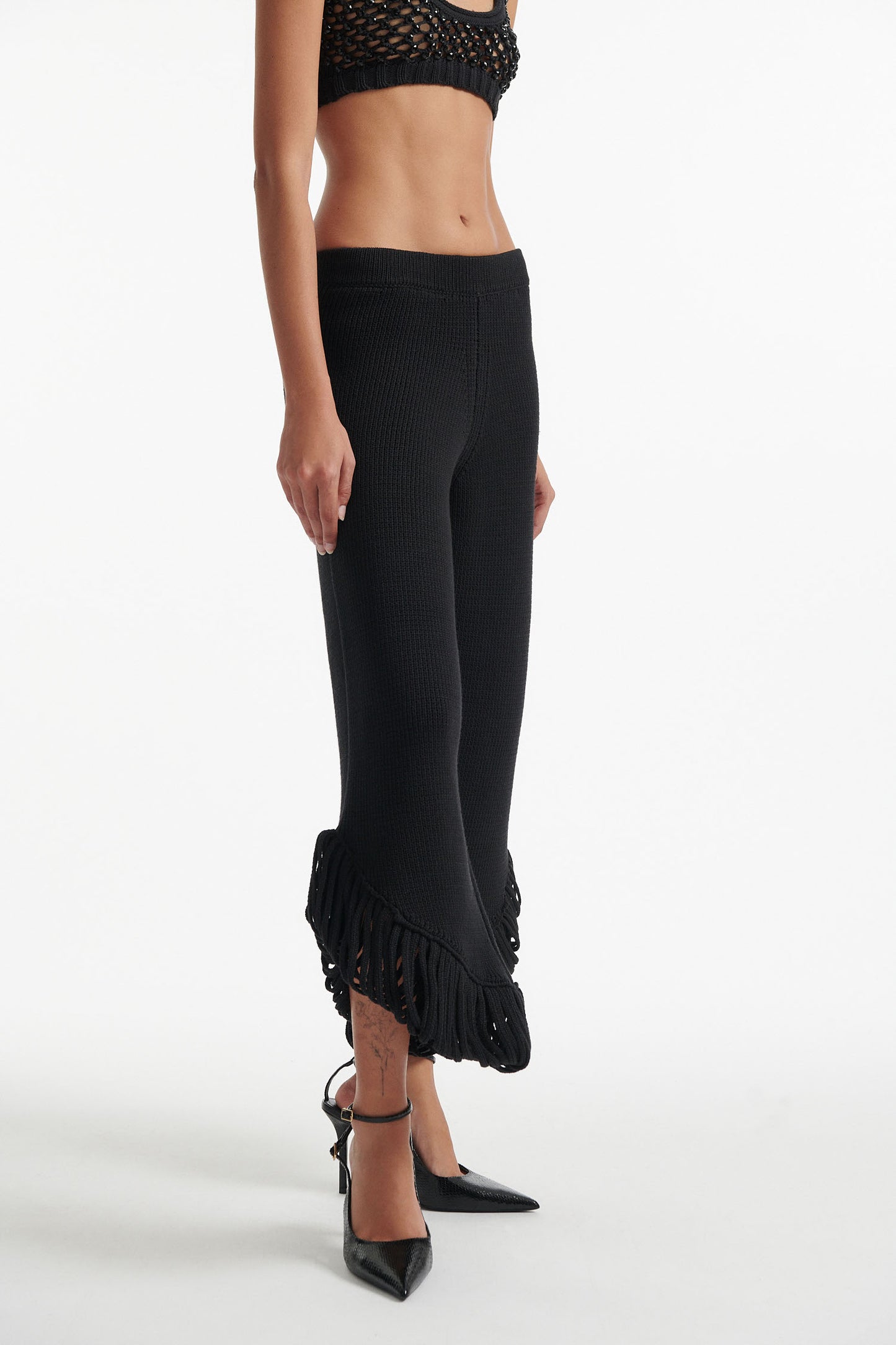 Female model wears short black knit trousers with fringe loops and black crystal embellished bralette with black high heels on white background.