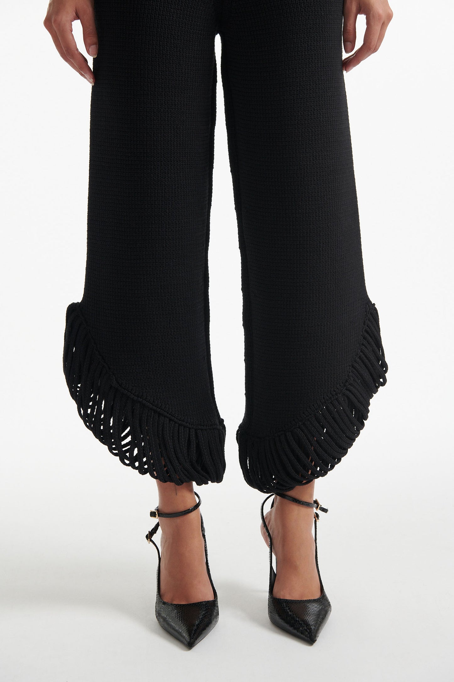 Female model wears short black knit trousers with fringe loops and black crystal embellished bralette with black high heels on white background.