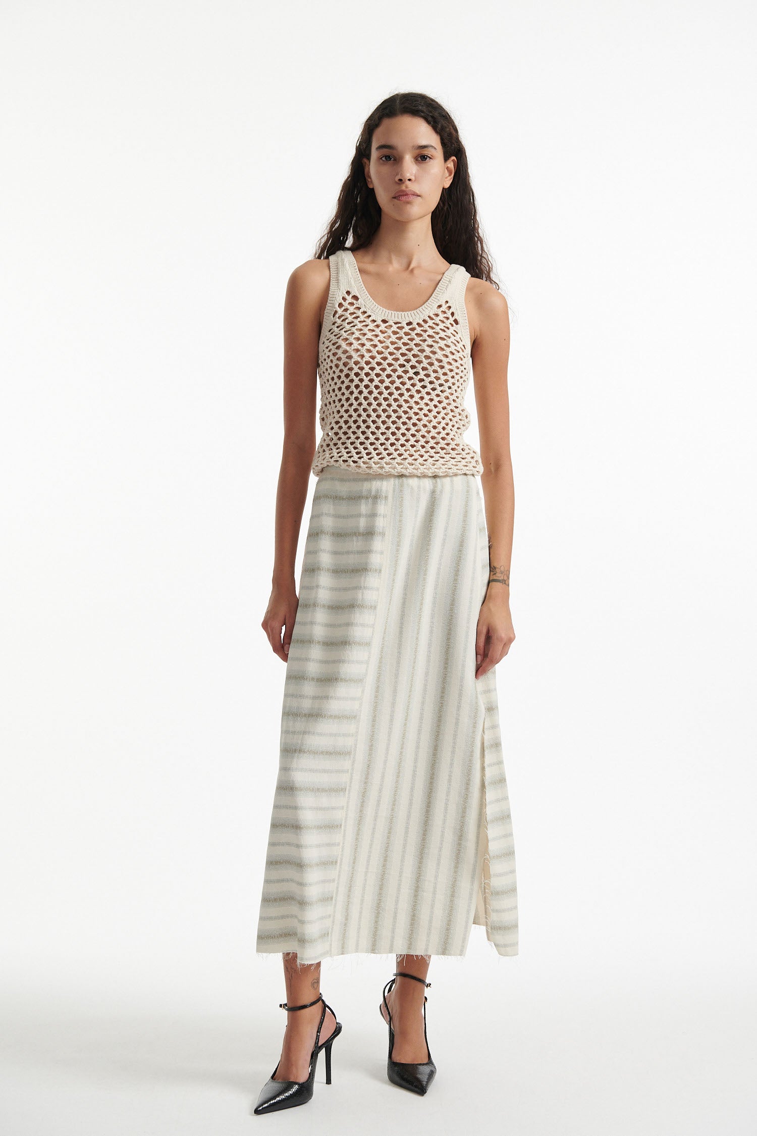 Female model wears beige mesh tank top and A line striped white skirt with black high heels on white background. 