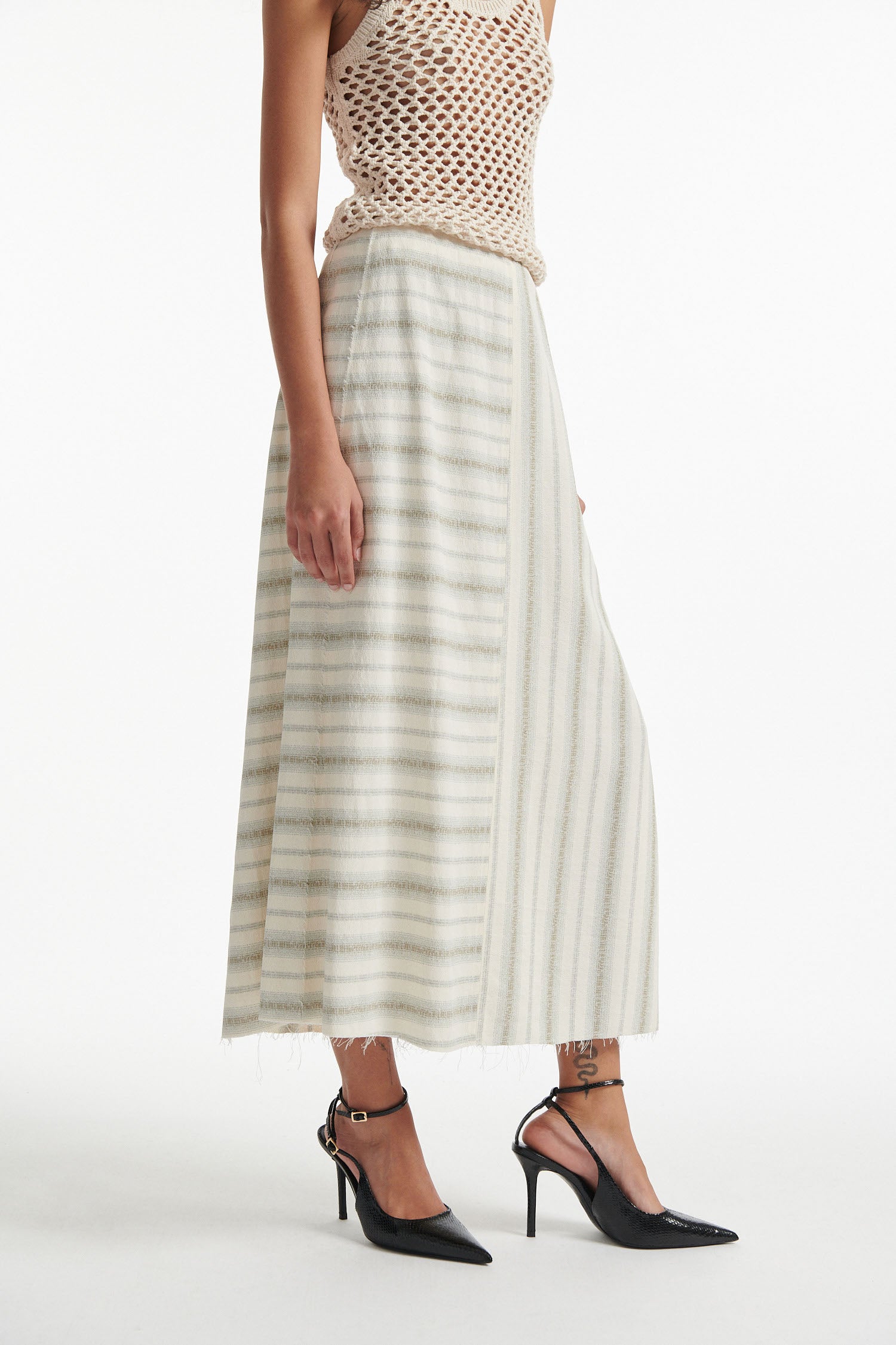 Female model wears beige mesh tank top and A line striped white skirt with black high heels on white background. 