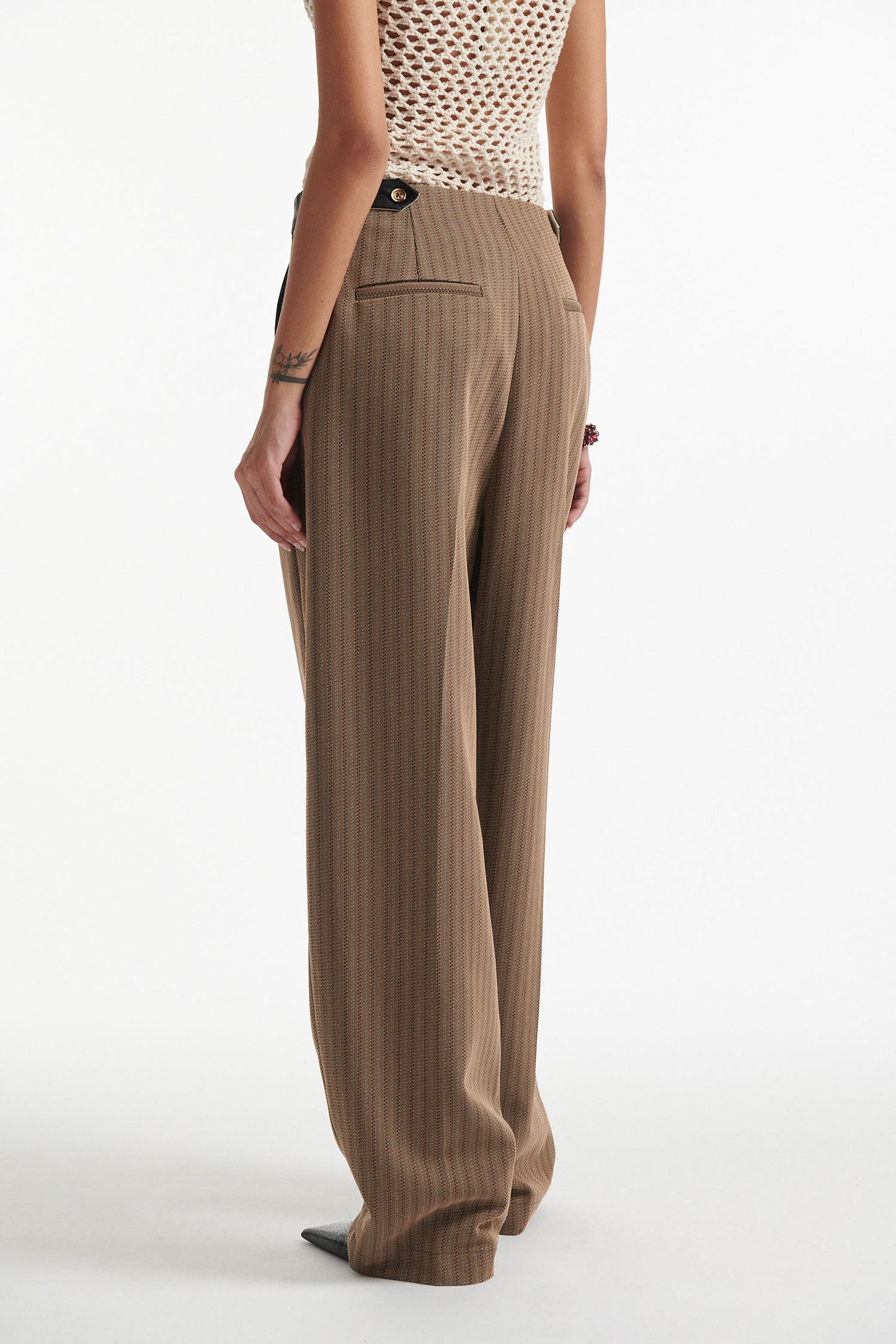 Female model wears beige cotton net tank top with brown pinstripe pleated trousers and black high heels on white background.