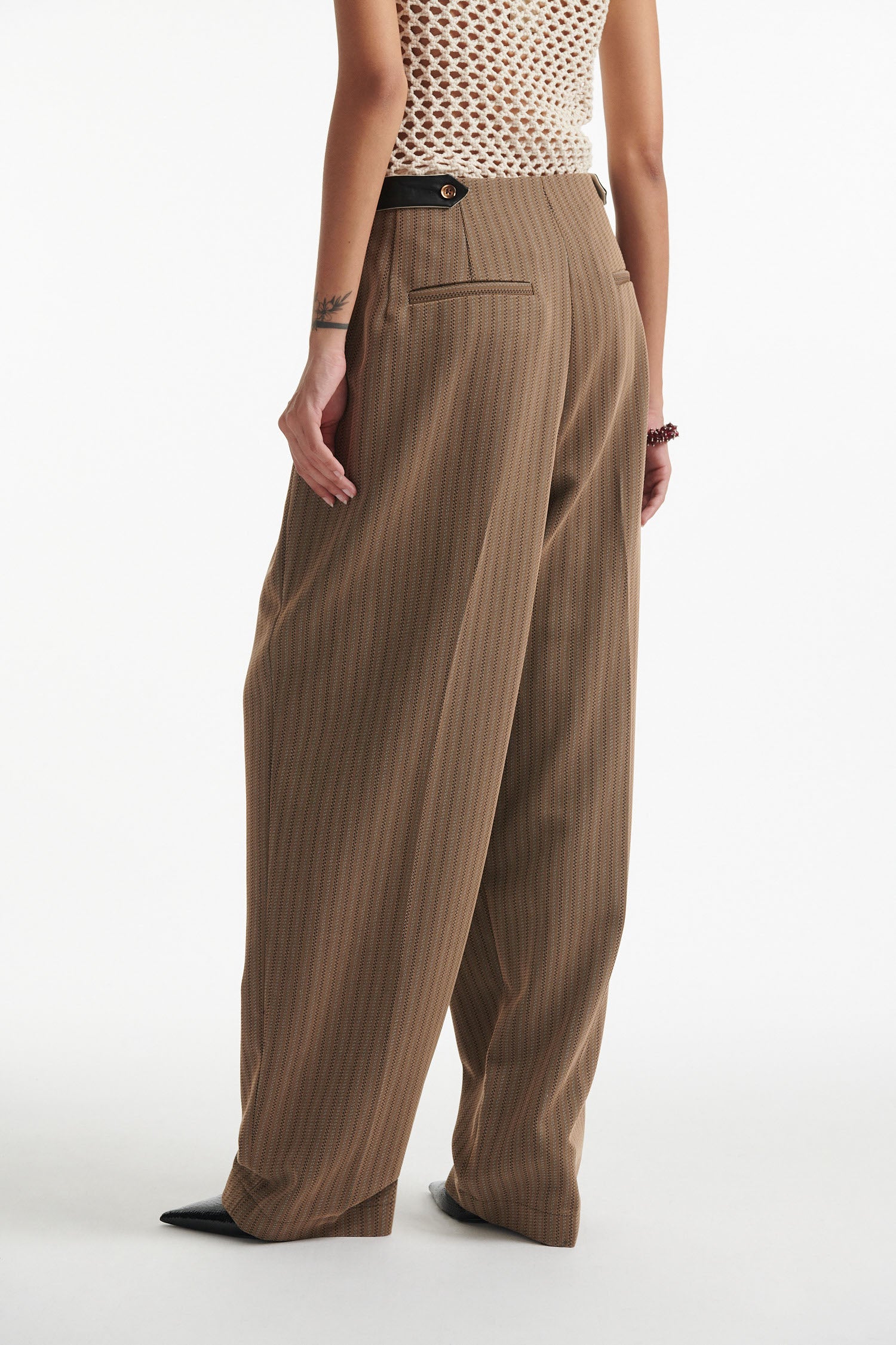 Female model wears beige cotton net tank top with brown pinstripe pleated trousers and black high heels on white background.