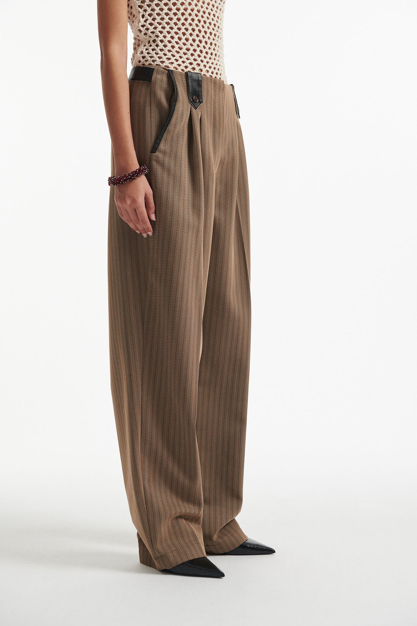 Female model wears beige cotton net tank top with brown pinstripe pleated trousers and black high heels on white background.