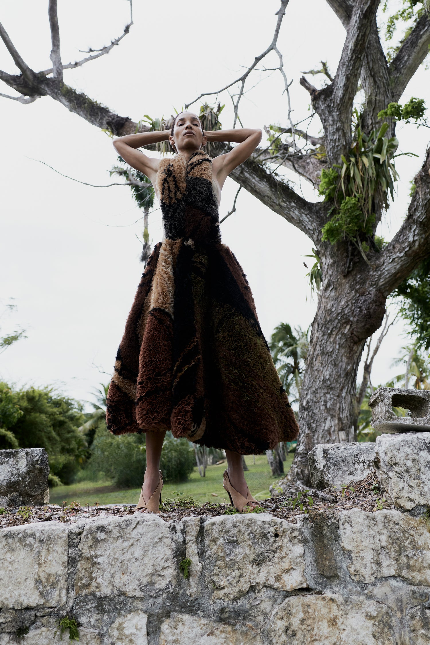 A woman stands outdoors on stone ruins beneath a large tree, wearing a voluminous patchwork dress made of dark brown, tan, and black textured fabrics.