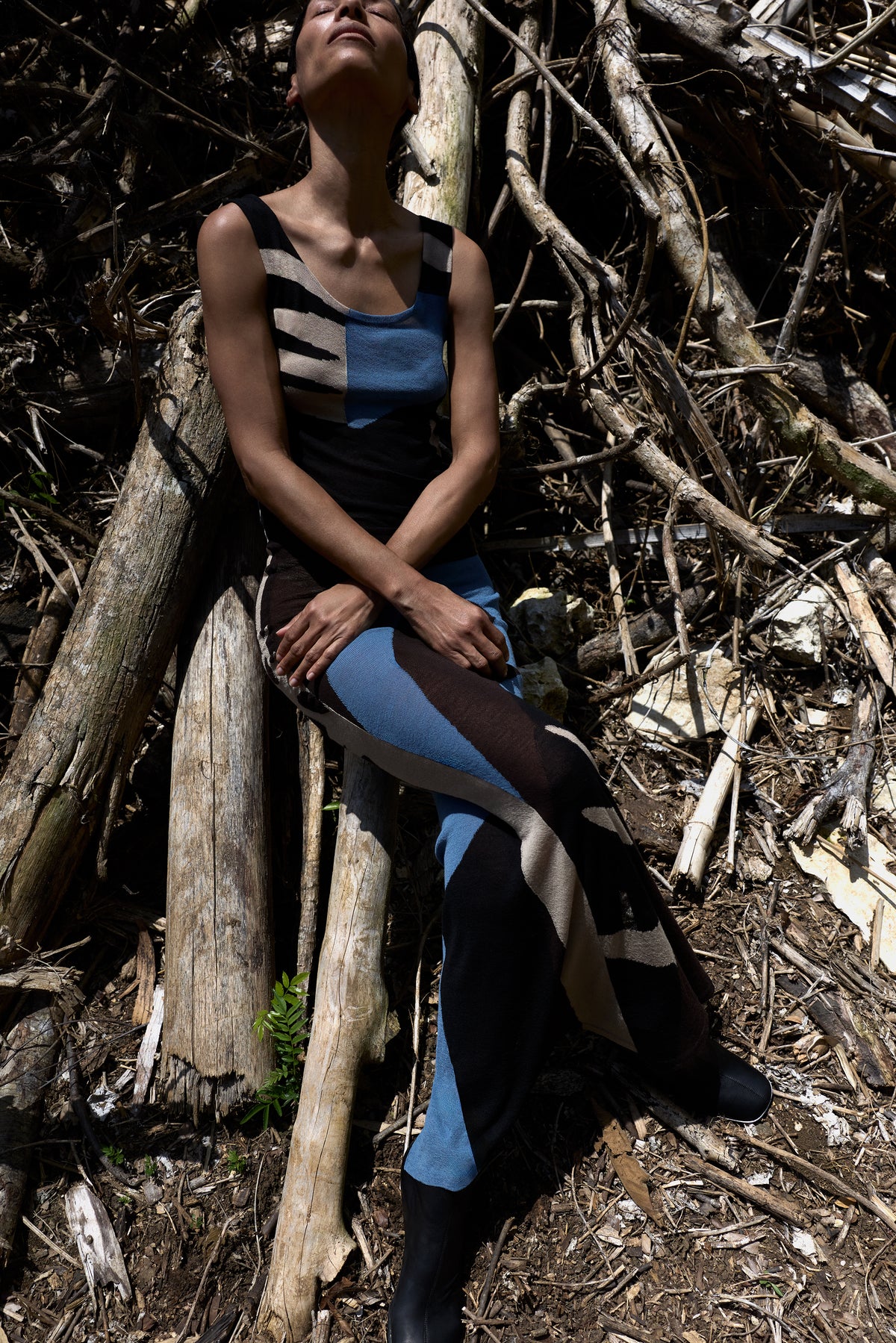 A model wearing a sleeveless color-blocked dress sits on logs in a forest clearing, surrounded by branches and tree trunks.
