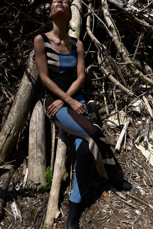 A model wearing a sleeveless color-blocked dress sits on logs in a forest clearing, surrounded by branches and tree trunks.
