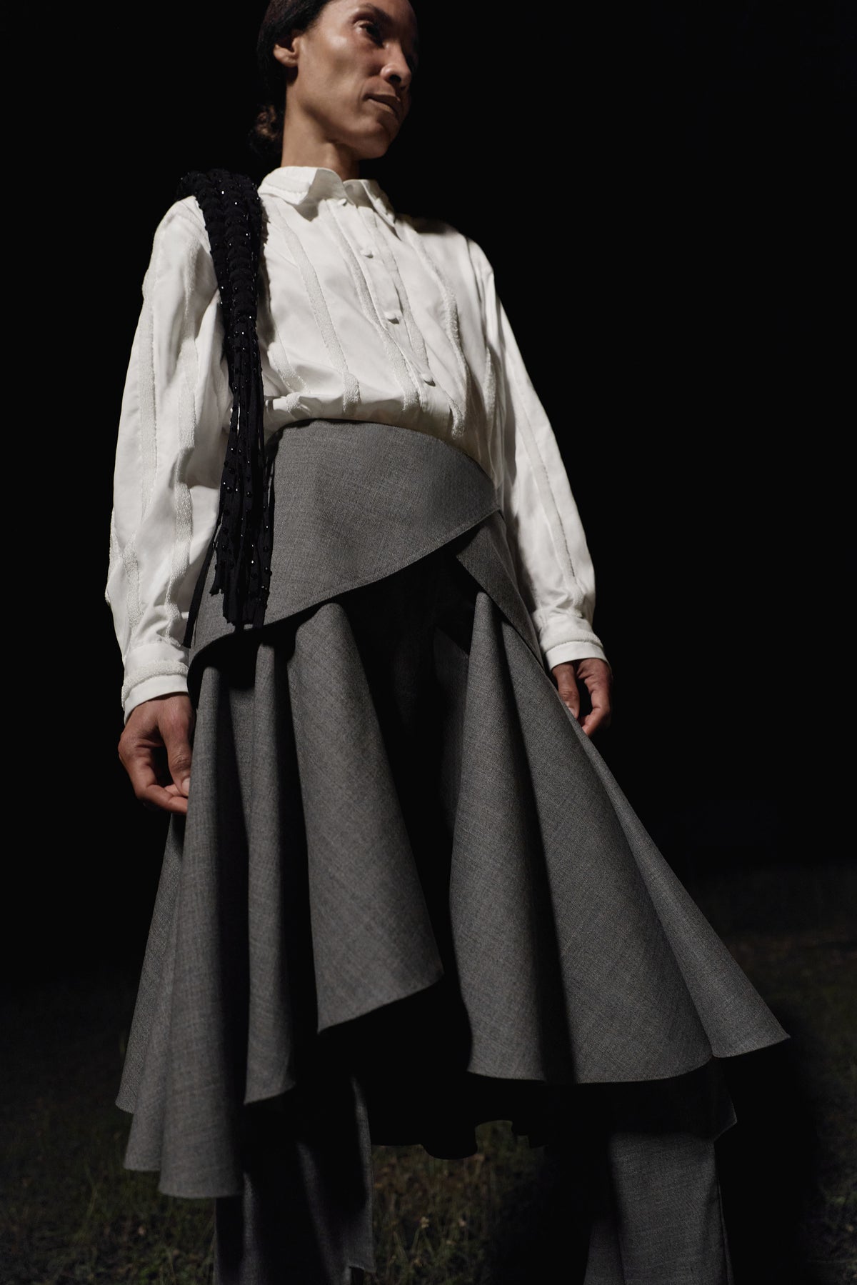 A model stands outside in the dark wearing a white button down shirt, grey skirt over trousers, and a black fringe embellishment on the shoulder