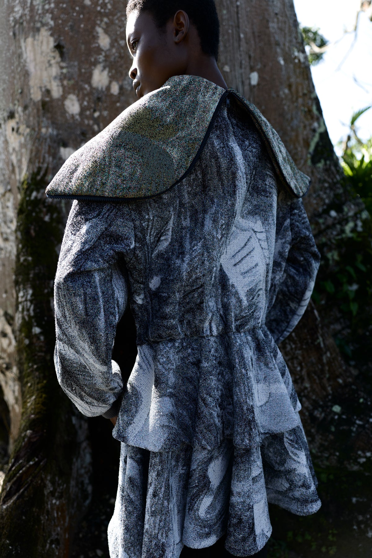 A model wearing a textured, painterly gray and black coat with a large structured shoulder cape, photographed outdoors against a tree trunk.