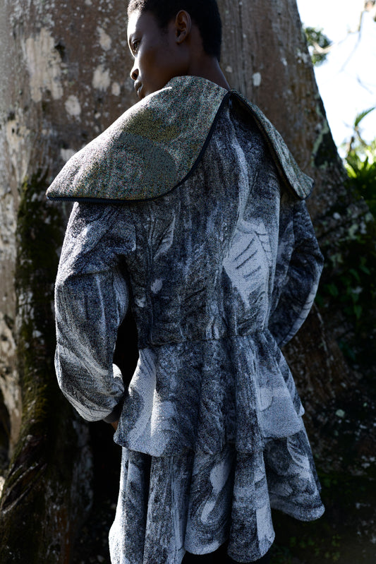 A model wearing a textured, painterly gray and black coat with a large structured shoulder cape, photographed outdoors against a tree trunk.