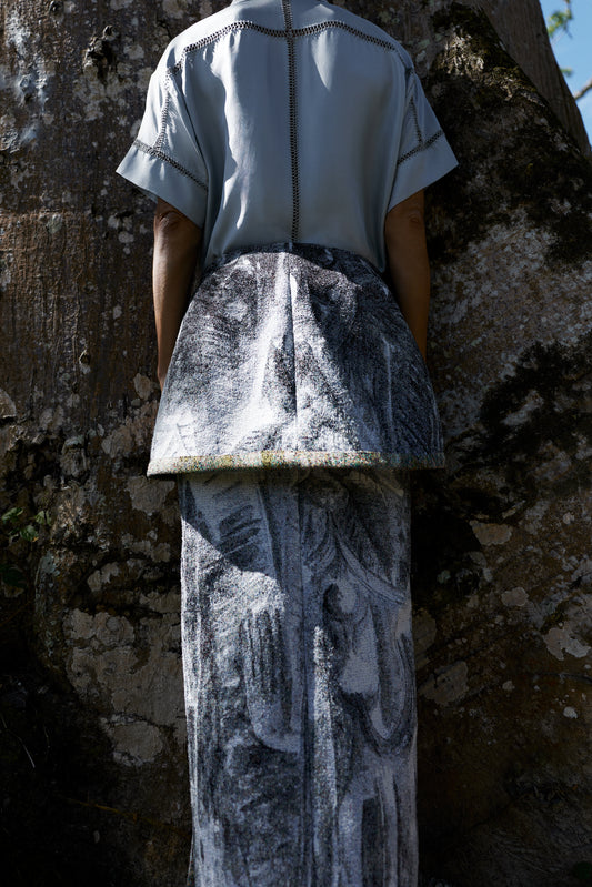 A model shown from behind wearing a light blue short-sleeve top and a long gray patterned skirt, standing outdoors against a large textured tree trunk.