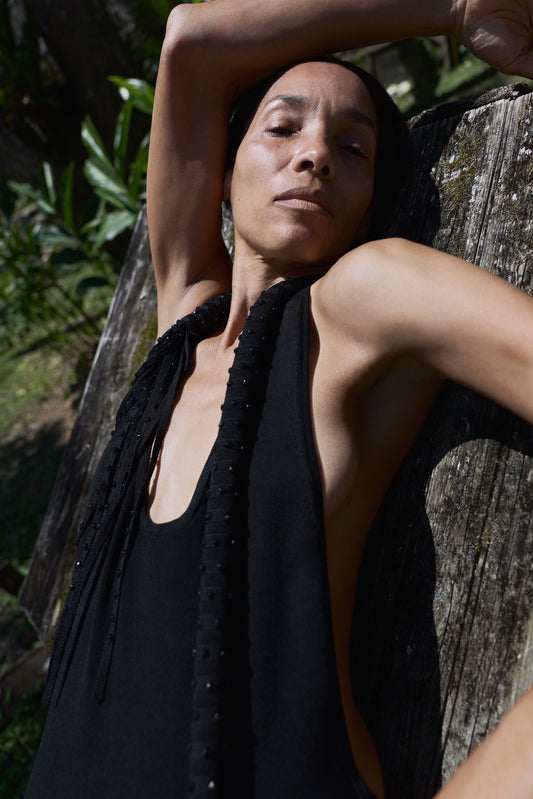 A woman wearing a black halter dress with a black fringed embellishment leans against a weathered wooden post outdoors, with green foliage visible in the background.