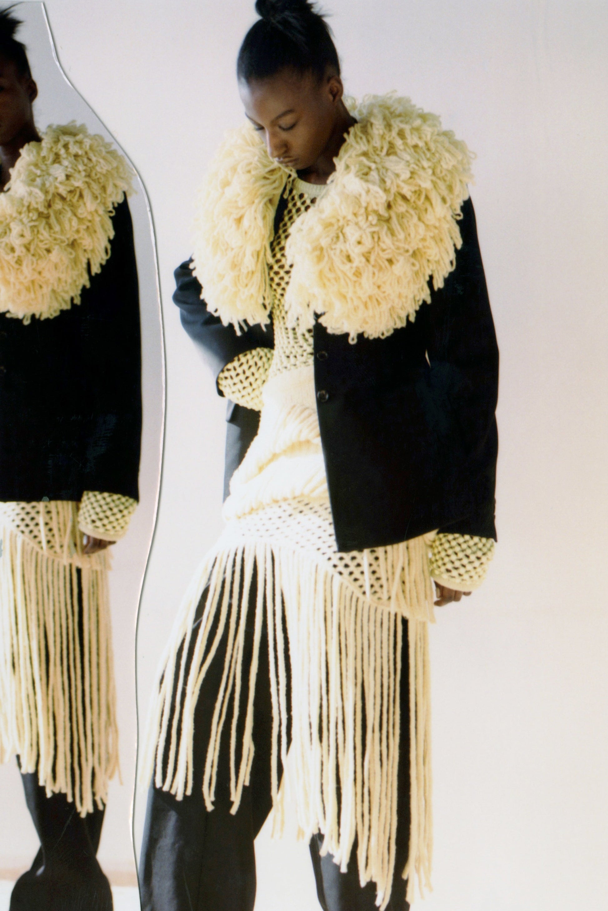 Model wearing a black and yellow outfit with crochet knit and fringe details, standing in front of a mirror photographed against a white background 
