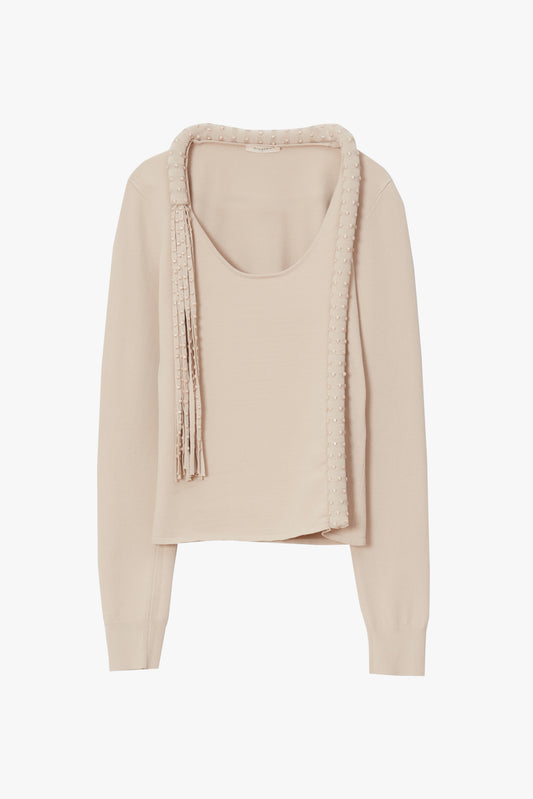 A light beige knit long-sleeve top with a scoop neckline, featuring a draped fringed trim detail across the chest, displayed on a clean white background.