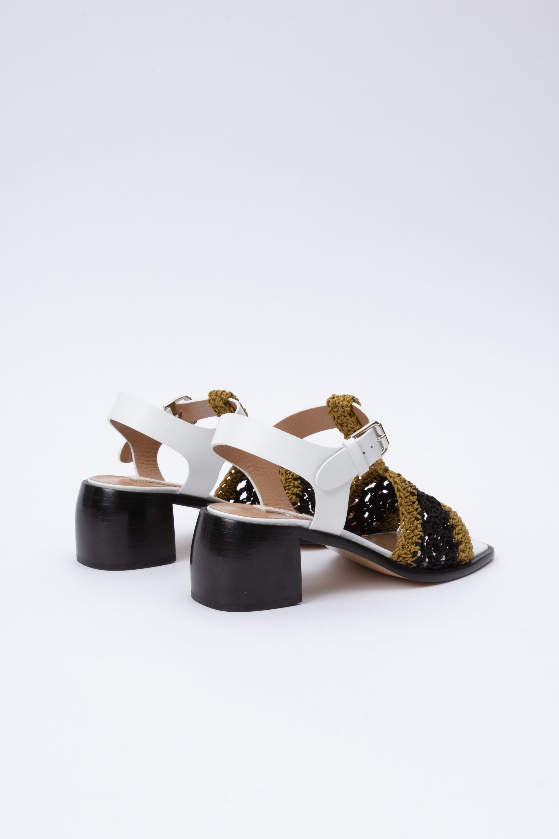 Open toe sandals with black leather base and white leather ankle straps. The block heel is slightly rounded and thick. Connected to the ankle strap is a toe strap made from hand-knit crochet in a cumin yellow and black. 