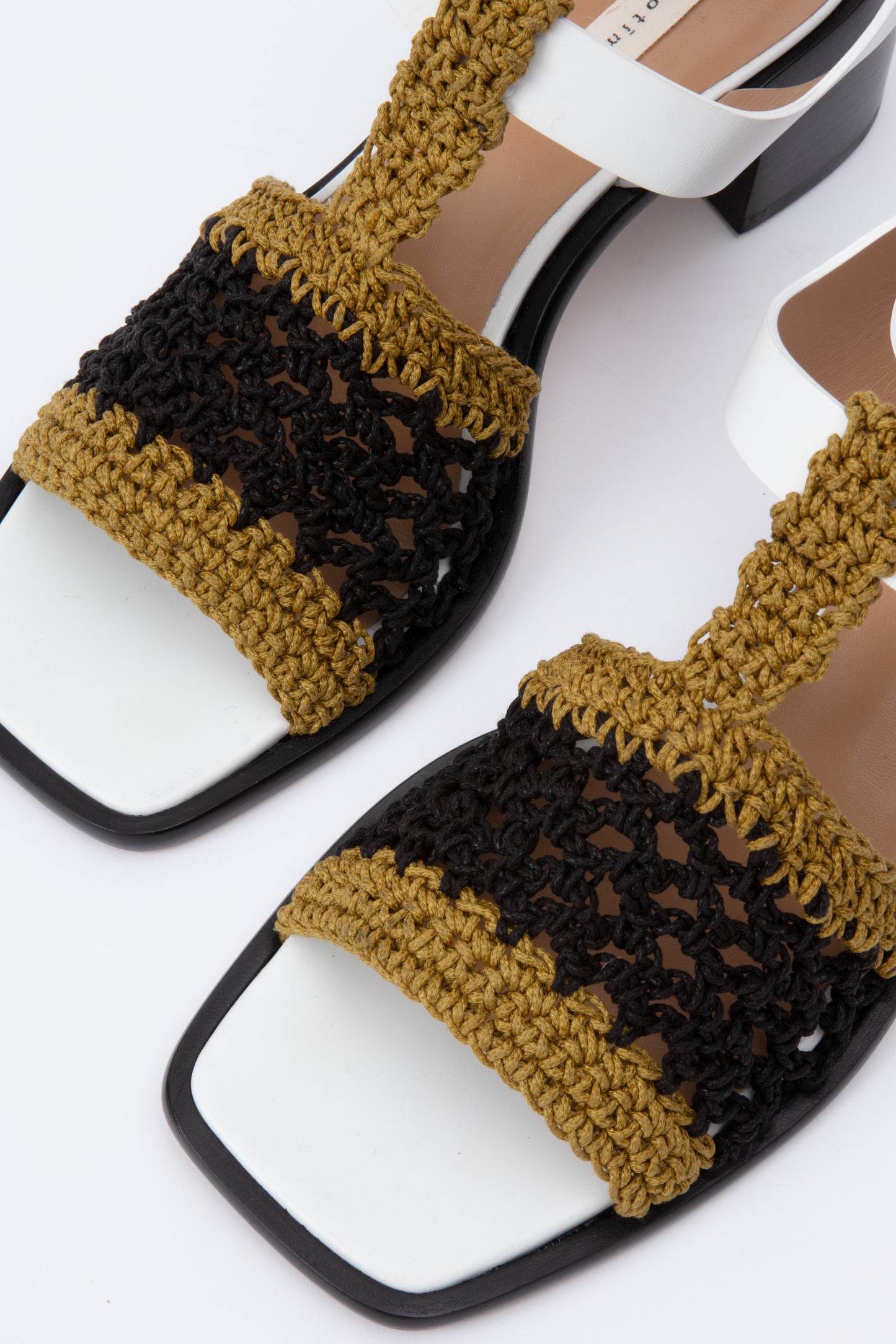 Open toe sandals with black leather base and white leather ankle straps. The block heel is slightly rounded and thick. Connected to the ankle strap is a toe strap made from hand-knit crochet in a cumin yellow and black. 
