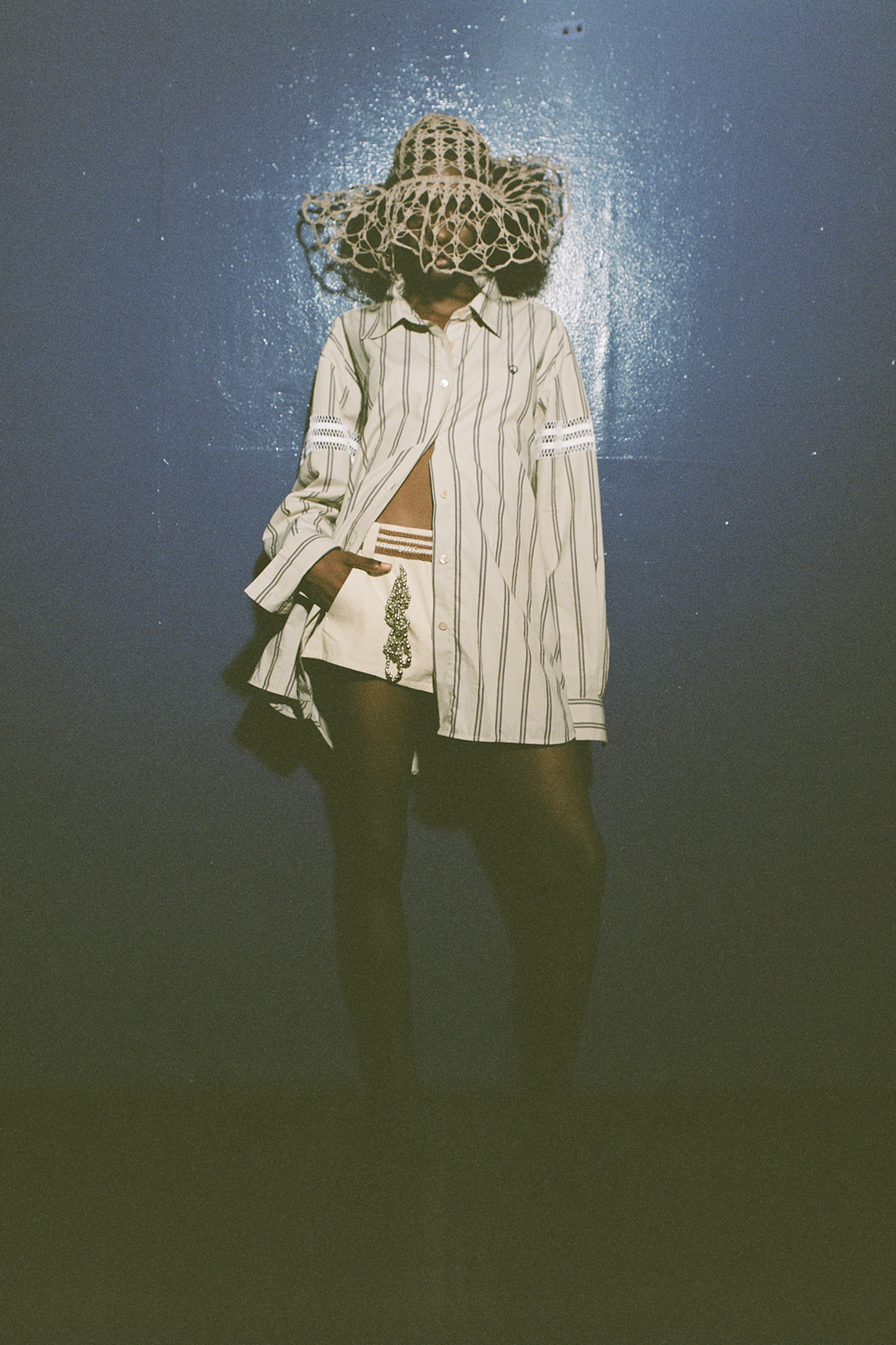 Model photographed on dark blue painted wall with flash. Model is wearing a vertical striped button down shirt in ecru-grey and canvas shorts with a brown crochet belt. The model's shirt has two vertical bands of white crochet on either arm. The model's shorts have two vertical front placed embellishments worked in silver spherical beads. The model is also wearing an ecru hand-crocheted hat.   