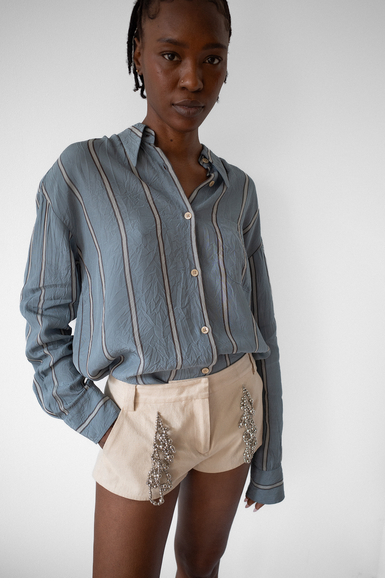 Model wears tan canvas short-shorts with two vertical front placed embellishments worked in silver spherical beads. Model also wears blue-grey viscose button down shirt.