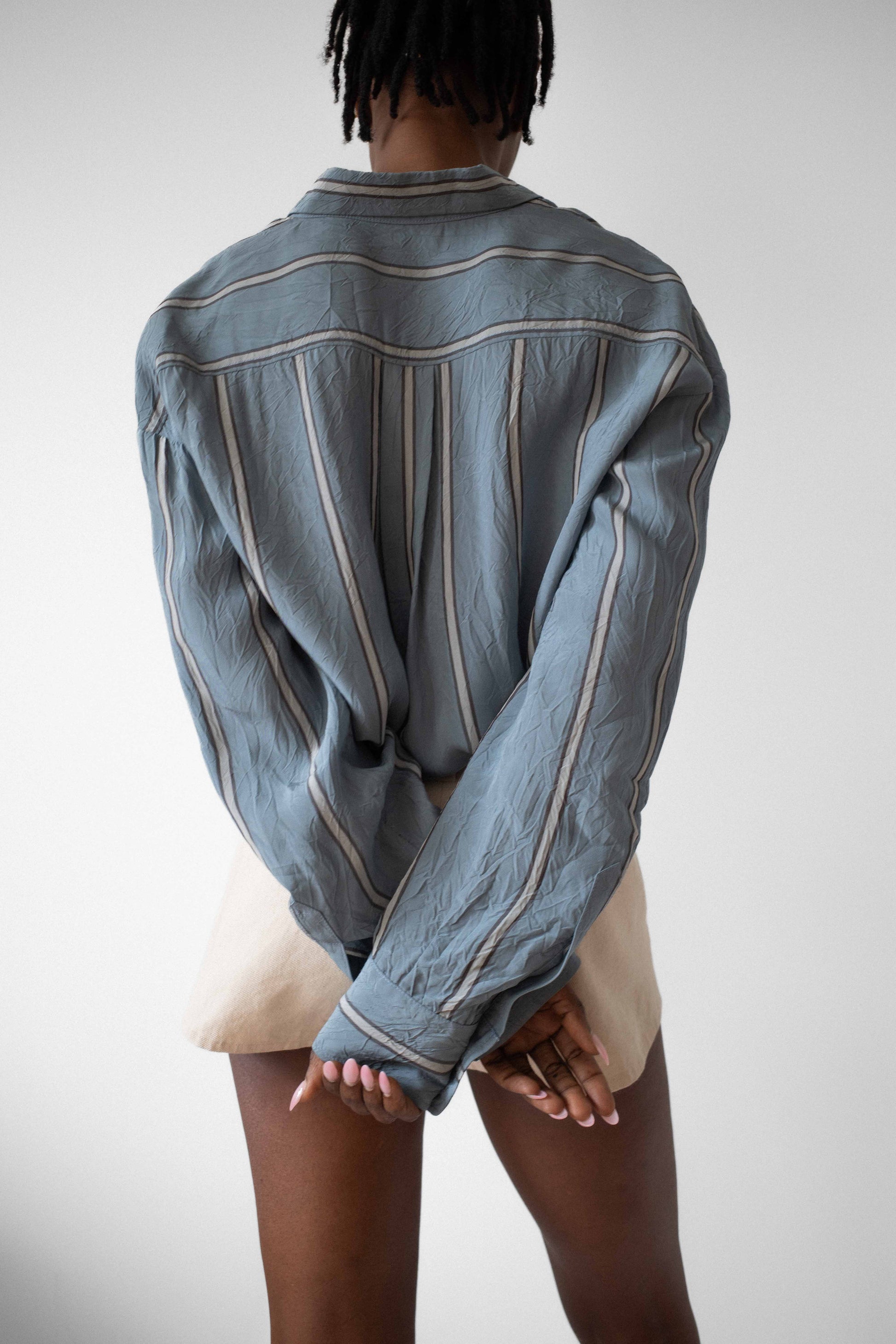 Model wears tan canvas short-shorts with two vertical front placed embellishments worked in silver spherical beads. Model also wears blue-grey viscose button down shirt.