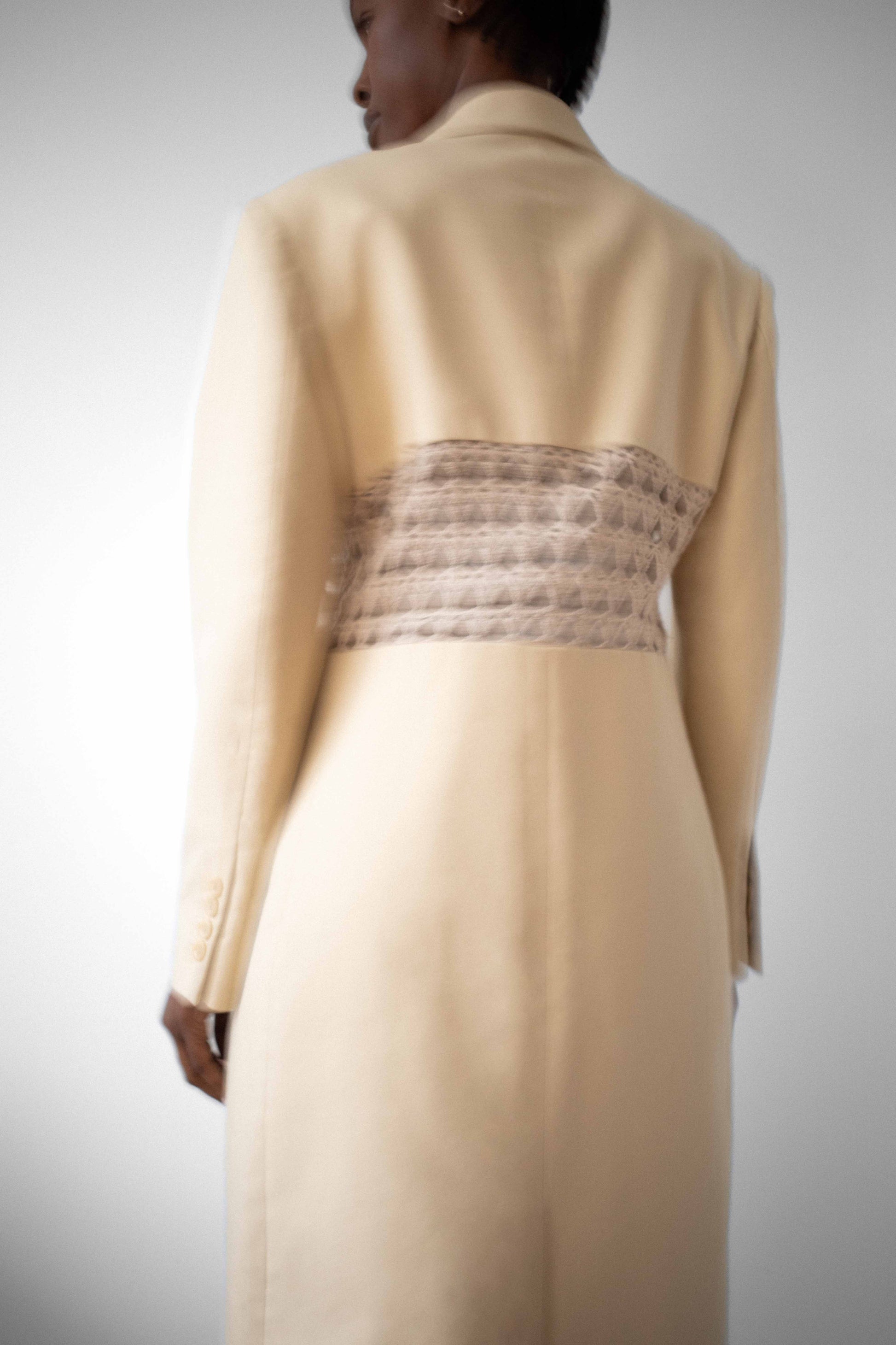 Blurry image of model wearing long beige structured coat with beige crochet paneling dividing the center back.