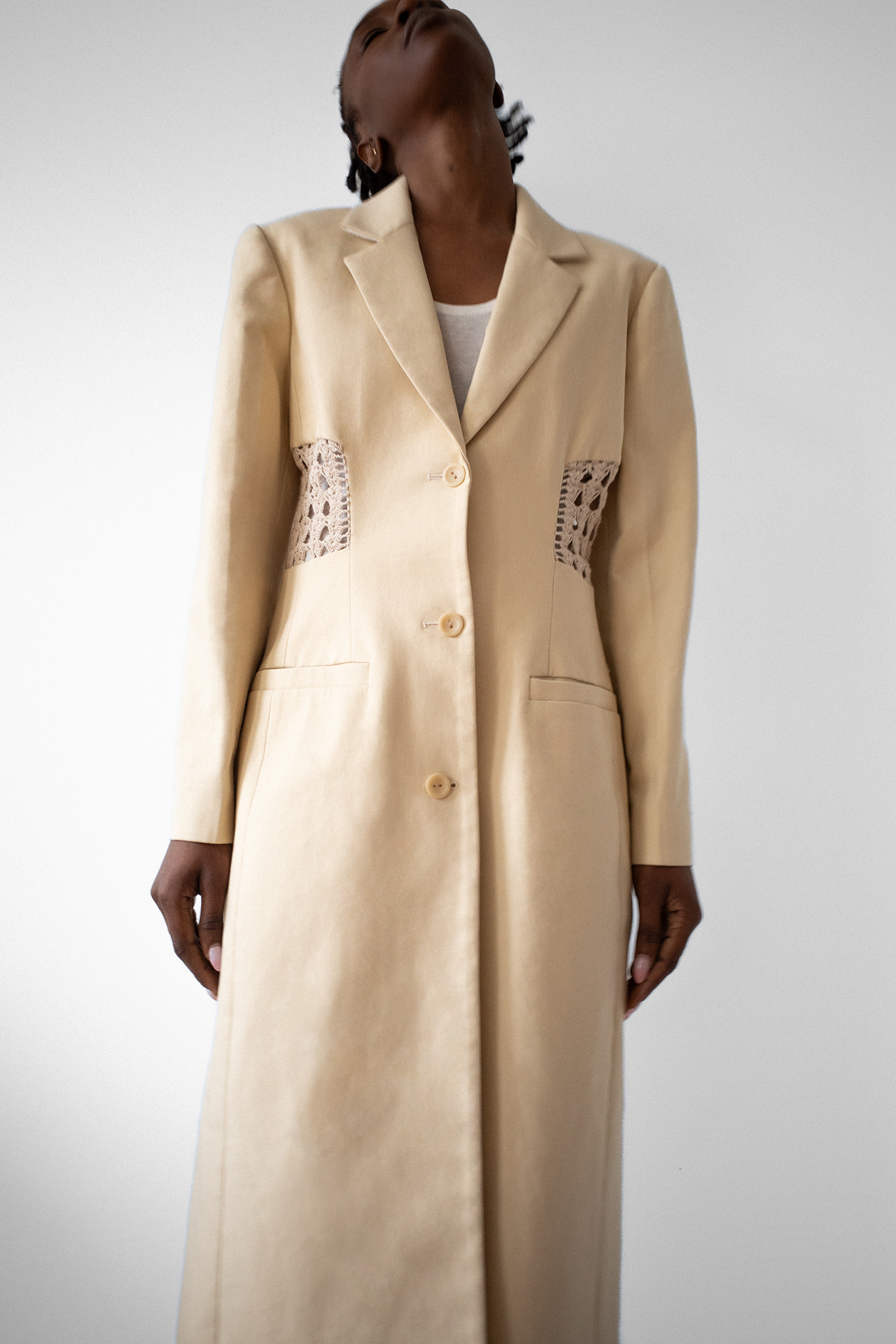 Model wearing long beige structured coat with beige crochet paneling dividing the center back. 