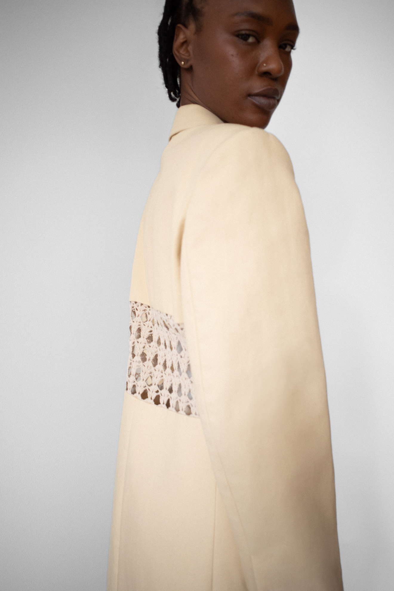 Model wearing long beige structured coat with beige crochet paneling dividing the center back. Model looks over her shoulder at the viewer.