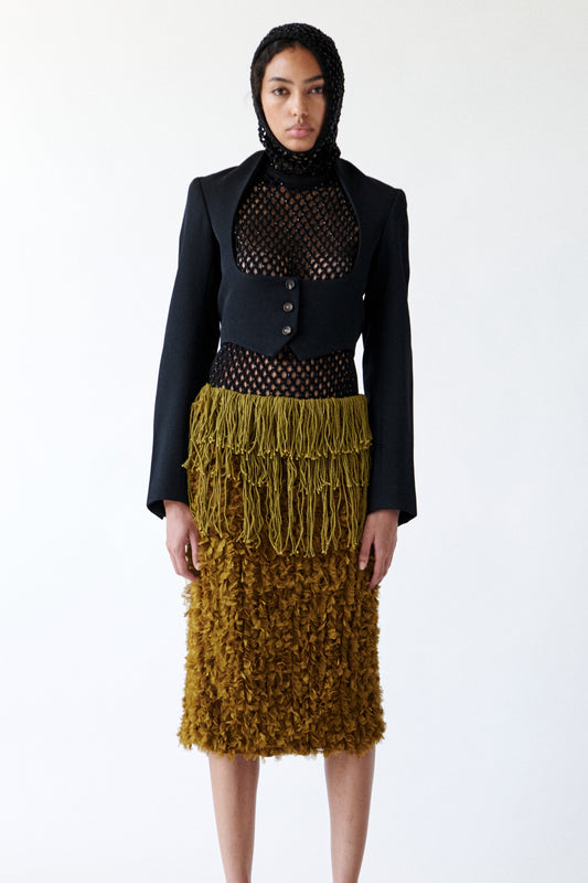 A model stands against a plain white studio background wearing a black tailored jacket over a black mesh top, paired with a textured olive-brown fluffy skirt with a braided drink yoke and matching sandals.