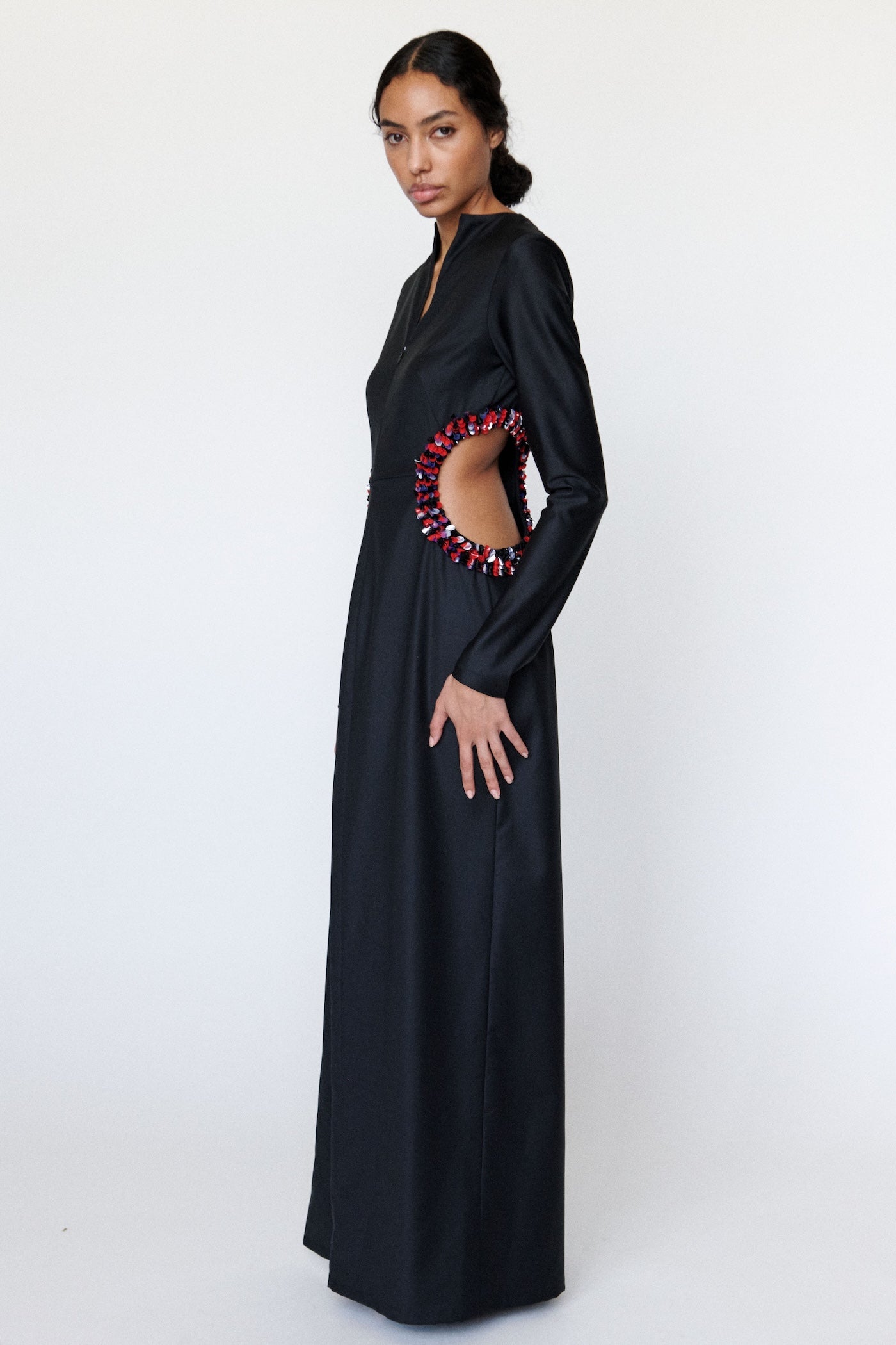 Model posing to the side is wearing a black dress with colorful circular cutout design. the cutout design is trimmed with a red, black, and silver palettes design on a white background 