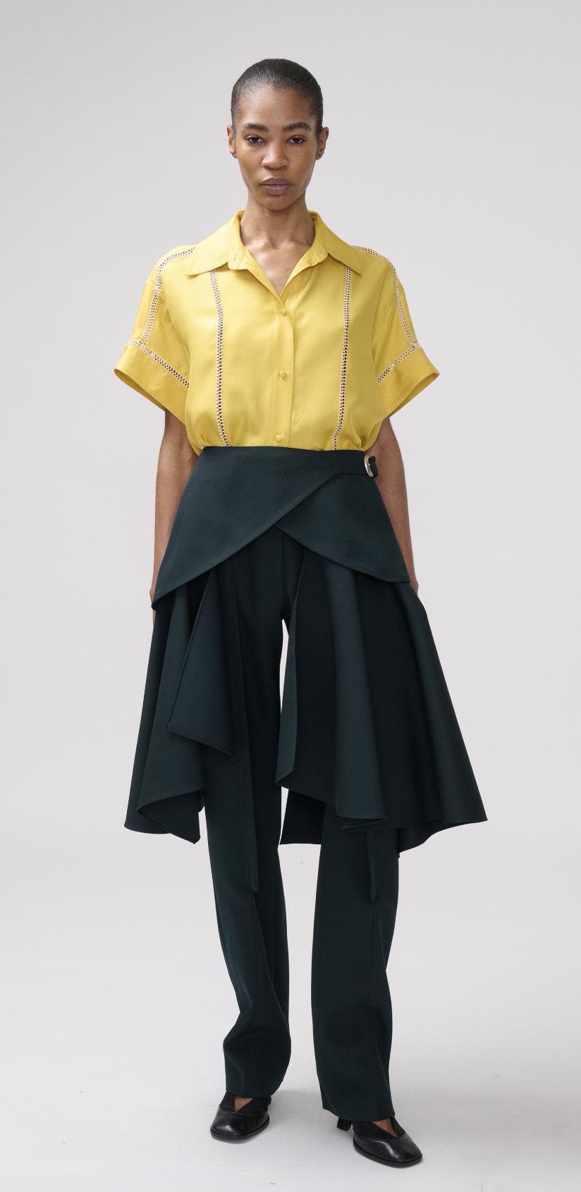 A model stands against a light gray studio background wearing a bright yellow short-sleeve button-up shirt and dark green wide-leg trousers.