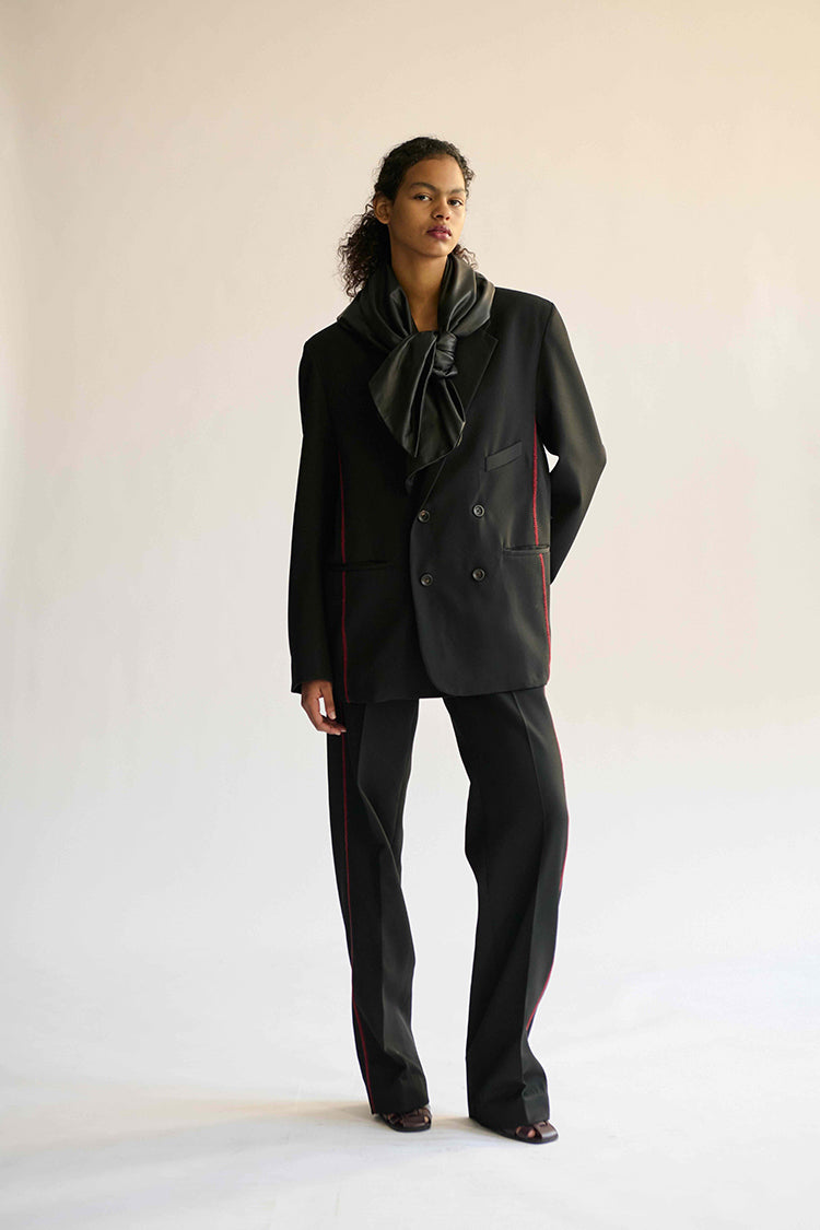 A model stands against a plain light beige studio background wearing a black double-breasted blazer with red stitching details, matching black trousers, and a dark gray scarf.