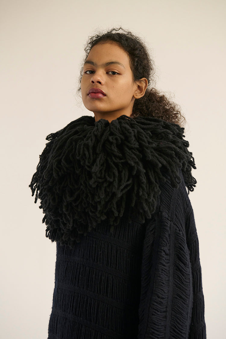 A model facing the camera, wearing a black textured knit sweater with a large, merino-wool knit collar.  