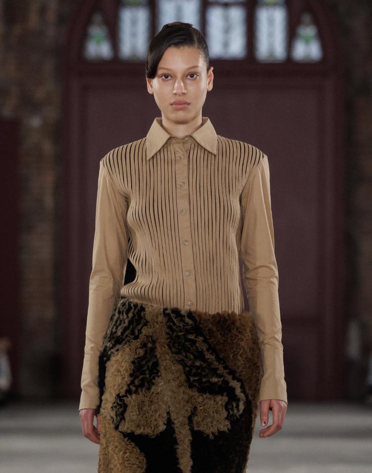 A model on the runway stands facing forward wearing a beige long-sleeve collared shirt with vertical pleats and a textured brown and black skirt.
