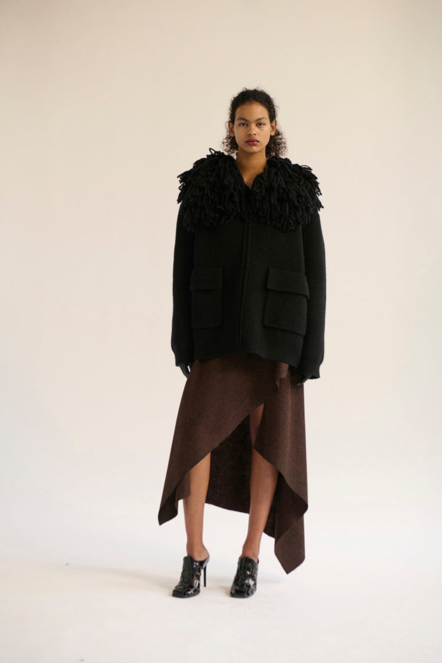 A model stands against a plain off-white studio background wearing a black cropped jacket with an oversized fringe collar and large front patch pockets.